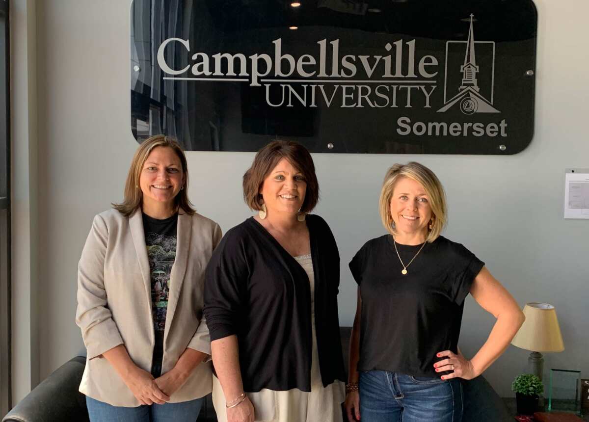 three women pose standing in and office under a Campbellsville University Somerset sign.