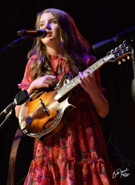 young girl with long hair and red flowered dress singing on stage with microphone playing mandolin