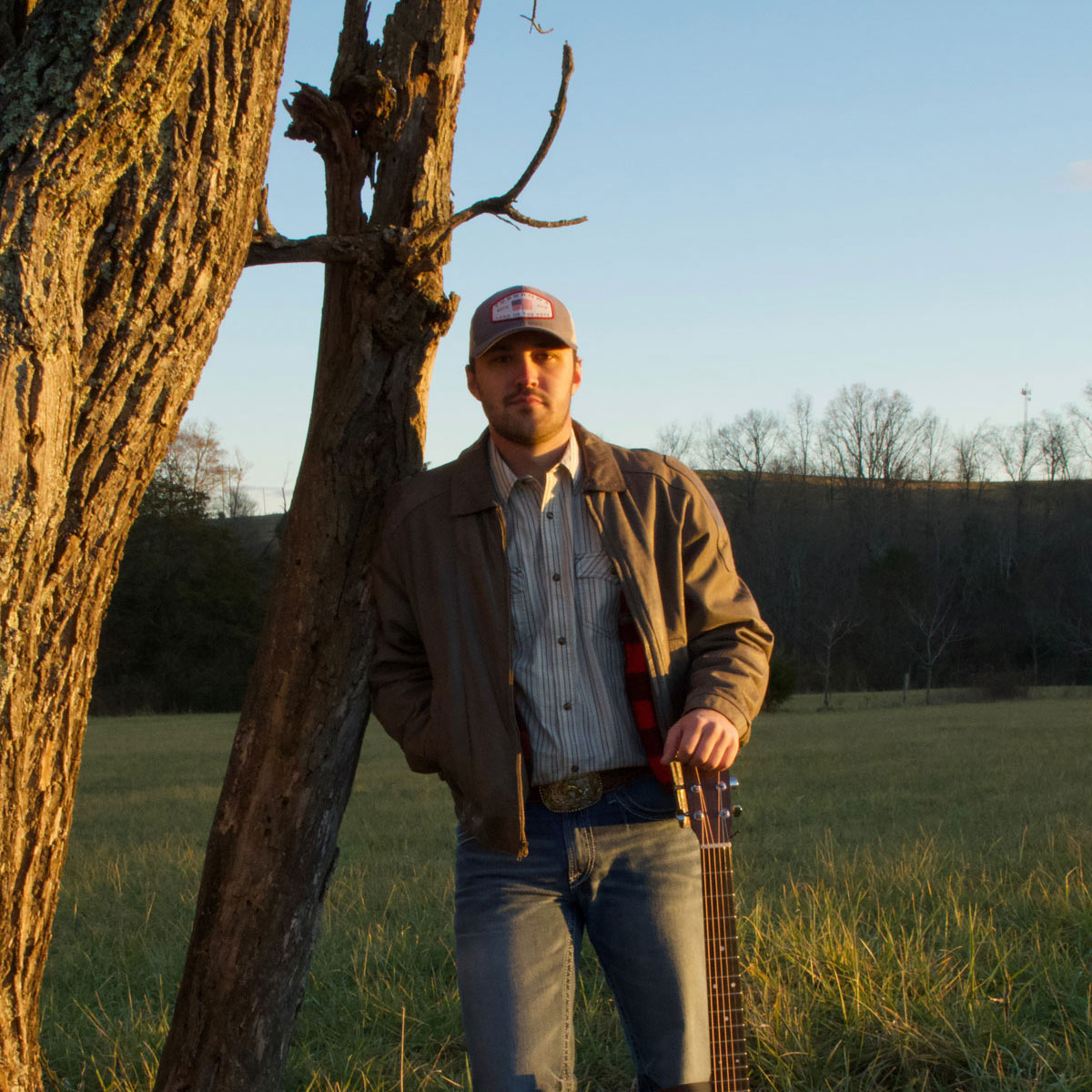 young man leaning against tree with guitar